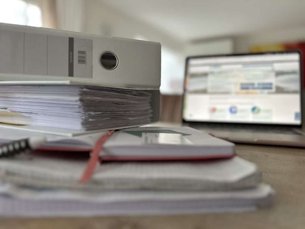 Versicherung zahlt nicht bei Wasserschaden? Wasserschaden wer zahlt? Tisch mit vielen Akten und Unterlagen, in Hintergrund Laptop mit Wasserschadenshilfe.de WDS Website geöffnet.